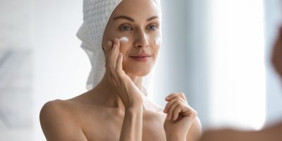 Attractive young adult woman applying facial cream looking in mirror